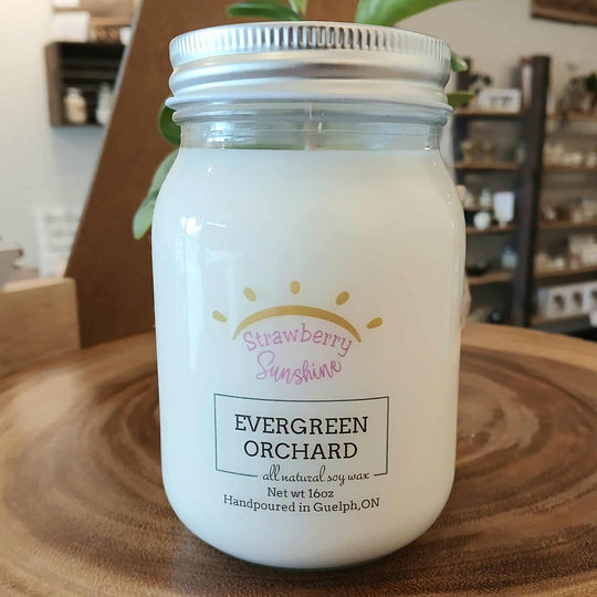 Candle in a glass jar labeled 'Strawberry Sunshine' by 'Evergreen Orchard' on a wooden surface.